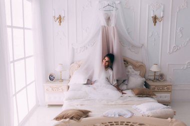 Güzel, mutlu, genç bir gelin yatak odasında gelinlikle poz veriyor. Gelinin özel sabahı..                     