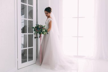 Gelinlik ve duvak giymiş genç ve güzel gelin içeride düğün çiçeği gibi poz veriyor.          