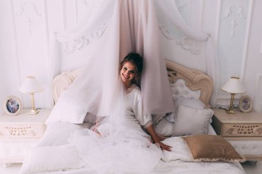 Güzel, mutlu, genç bir gelin yatak odasında gelinlikle poz veriyor. Gelinin özel sabahı..                     