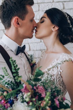 Mutlu genç gelin ve damat, tuğla duvarın yanında düğün buketi ile duruyorlar.       