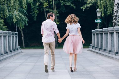 Genç mutlu çiftin el ele tutuşup yazın parkta yürüyüşünün arka görüntüsü.