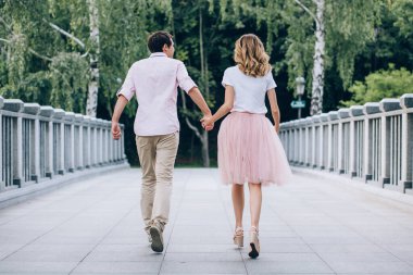 Yaz parkında köprüde el ele tutuşan genç bir çiftin arka görüntüsü.