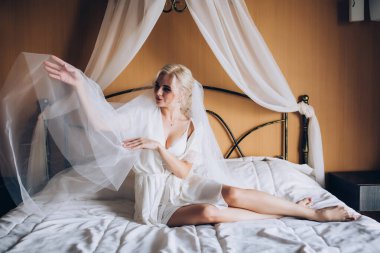 Genç, güzel sarışın gelin otel odasında yatakta poz veriyor.