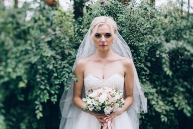 Güzel genç gelin elinde buketle parkta poz veriyor.              