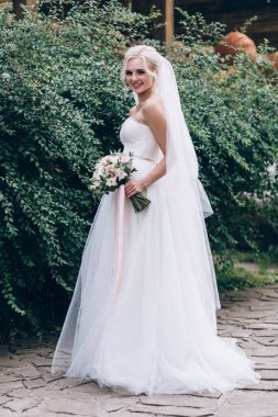 Güzel genç gelin elinde buketle parkta poz veriyor.              