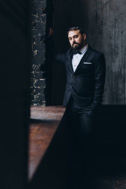 Siyah duvarın yanında poz veren takım elbiseli ve papyonlu yakışıklı genç adam.