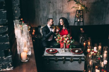 Şık, genç aşıklar ellerinde bardaklarla kanepede oturup mum yakıyorlar.      