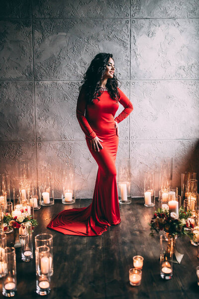 красивая женщина в красном платье позирует в комнате со свечами на полу            
