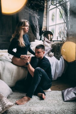 Sıcak bir odada, yatağın yanında oturan mutlu bir çift, erkek, kadın yerde otururken kadının bacaklarına nazikçe sarılır.