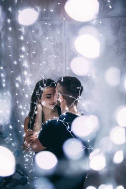 Gri duvarın yanındaki stüdyoda poz veren genç ve güzel bir çift, Bulanık çelenkler ve yeni yıl ışıkları.