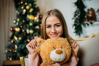 Örgü kazaklı genç bir kadın elinde oyuncak ayıyla bir Noel ağacının yanında oturuyor.  