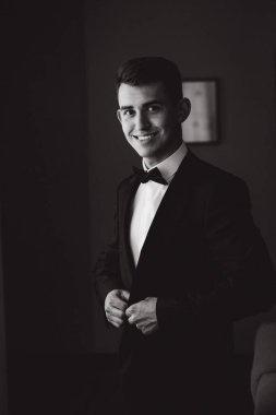 Smokinli genç bir adamın kameraya gülümseyişinin siyah beyaz görüntüsü.