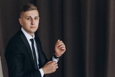 Siyah takım elbiseli, kravatlı ve beyaz gömlekli genç bir adam kameraya bakıyor.