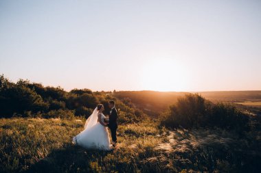 Güzel genç gelin ve yakışıklı damat gün batımında sahada poz veriyorlar. 