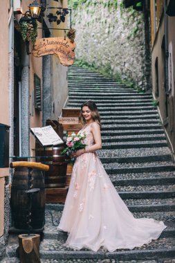 Genç gelin, elinde buketle eski bir kasabanın merdivenlerinde poz veriyor.