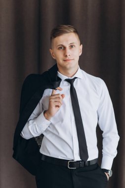 Stüdyoda poz verip kameraya gülümseyen şık kıyafetli genç bir adam.