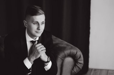 Takım elbiseli genç adam sandalyede oturuyor ve yan bakıyor, siyah beyaz.