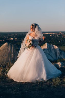 Gün batımında dağlarda elinde bir buket çiçekle beyaz elbiseli güzel bir gelin.