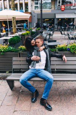 Genç bir kadın şehirde bankta oturan erkek arkadaşına sarılıyor.