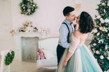 Şık elbiseli mutlu genç çift Noel ağacının yanında sarılıyorlar.  