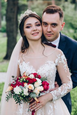 Gelin ve damat parkta yeşil ağaçların arka planında sarılıyorlar.