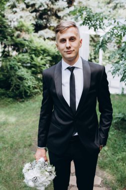 Yakışıklı damat düğün buketini tutuyor ve dışarıda kameraya gülümsüyor.