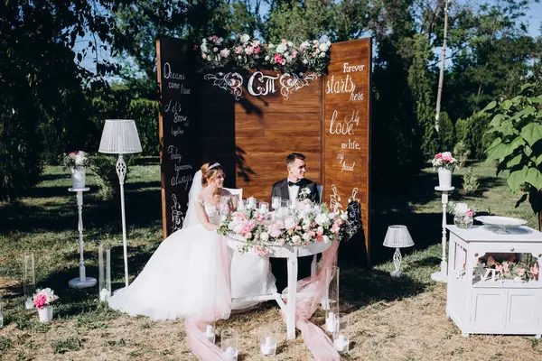 Güzel, mutlu gelin ve damat açık hava düğün töreninde ellerinde mumlar ve çiçeklerle masada oturuyorlar..              
