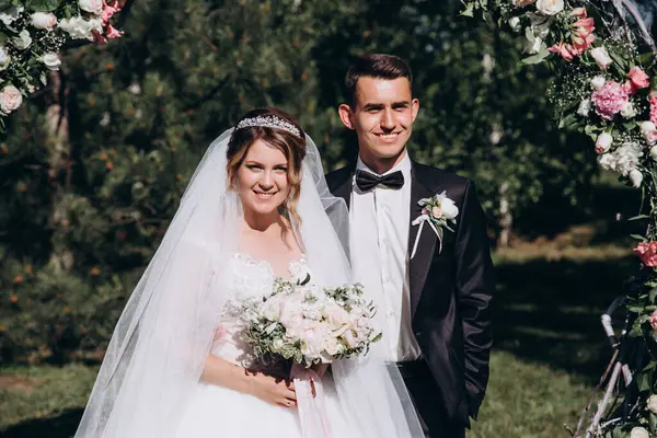 Güzel, mutlu gelin ve damat, düğün günlerinde çiçek kemerinin yanında.    