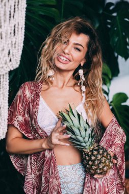 Mutlu sarışın kadın yeşil bitkilerle süslenmiş rahat stüdyoda egzotik ananasla poz veren iç çamaşırı ve kırmızı eşarp giyiyor. 