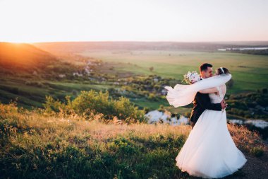 Mutlu gelin ve damat gün batımında dağlarda sarılıyor.