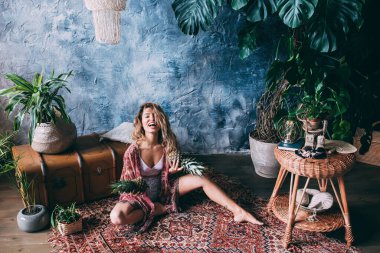 Yerde oturan kadın, yeşil bitkilerle süslenmiş rahat stüdyoda egzotik ananaslarla poz veriyor. 