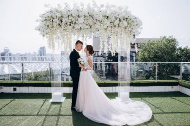 Birbirine aşık düğün çifti beyaz çiçeklerle süslenmiş güzel kemerin yanında yan yana duruyor. 