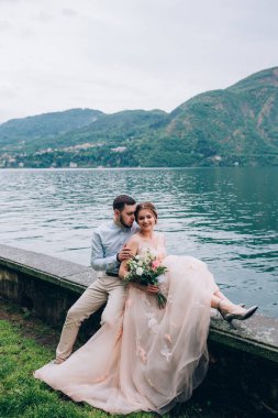 Düğün çifti dağların ve gölün arka planında oturup sarılıyorlar. 