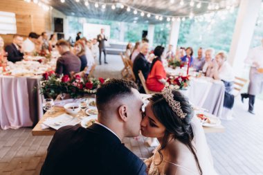 Genç bir çift masada oturuyor ve düğün resepsiyonunda öpüşüyor.