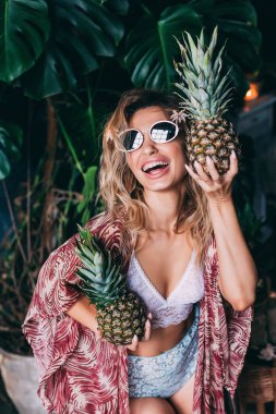 Güneş gözlüklü, egzotik ananaslarla poz veren genç ve mutlu bir kadın yeşil bitkilerle süslenmiş rahat bir stüdyoda. 