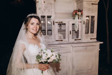 Beyaz elbiseli güzel gelin elinde düğün buketi ve gece parkta düğün dekoruyla dikiliyor.