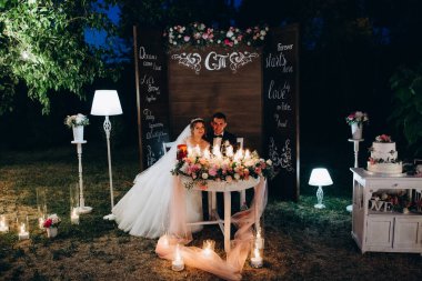Gelin ve damat geceleri yanan mumların ve açık hava düğün dekorunun yanında oturuyorlar.