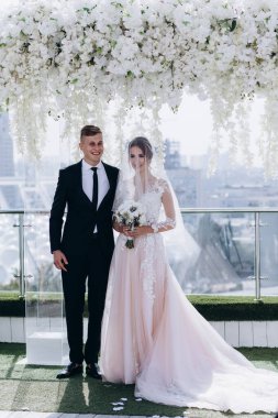 Gelin ve damat, beyaz çiçekli gelinlik içinde dekoratif çiçek kemerinin yanında duruyorlar.