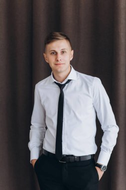 Beyaz gömlekli genç adam elleri ceplerinde kameraya gülümsüyor.