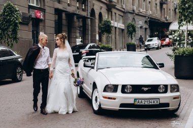 Güzel genç bir çift gelin ve damat şehirde beyaz düğün arabasının yanında poz veriyorlar.