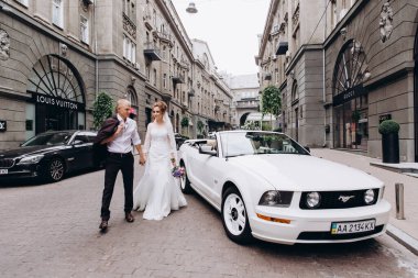 Güzel genç bir çift gelin ve damat şehirde beyaz düğün arabasının yanında poz veriyorlar.