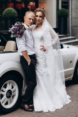 Güzel genç bir çift gelin ve damat şehirde beyaz düğün arabasının yanında poz veriyorlar.