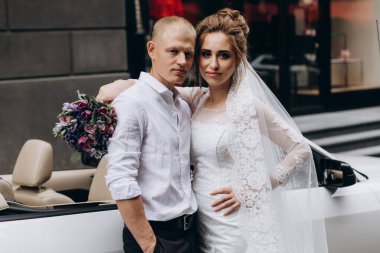 Güzel genç bir çift gelin ve damat şehirde beyaz düğün arabasının yanında poz veriyorlar.