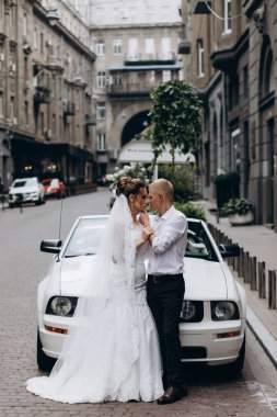 Güzel genç bir çift gelin ve damat şehirde beyaz düğün arabasının yanında poz veriyorlar.