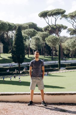 Tişörtlü ve şortlu bir adam park arka planında poz veriyor. 