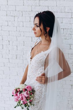 Beyaz elbiseli ve peçeli güzel gelinin portresi beyaz tuğla duvarda çiçek demeti ile poz veriyor.             
