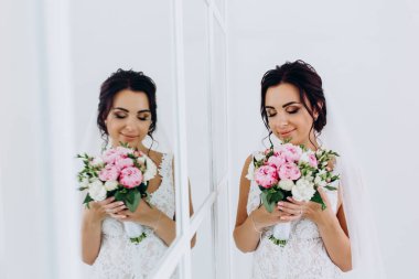 Beyaz elbiseli güzel genç gelin zarif iç kısımda düğün buketi ile poz veriyor.          