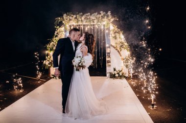 Mutlu gelin ve damat parkta düğün kemerinin yanında poz veriyor.          