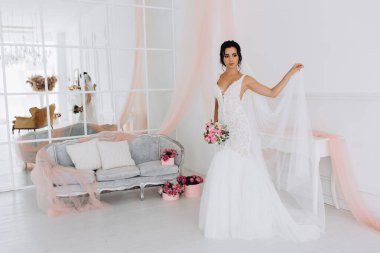 Beyaz elbiseli güzel genç gelin zarif iç kısımda düğün buketi ile poz veriyor.          