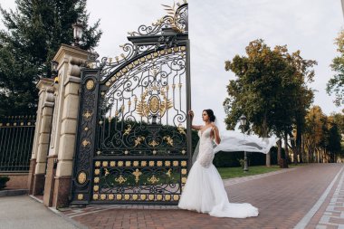 Beyaz gelinlik ve duvak içinde genç ve güzel bir gelin parkın kapısında poz veriyor.       